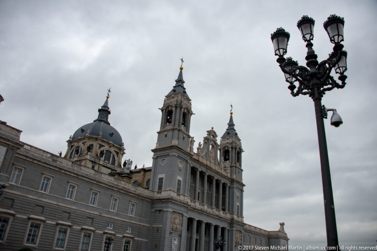 Almudena Cathedral dome and towers rising beside the palace precinct, with an ornate street lamp against a cloudy sky.