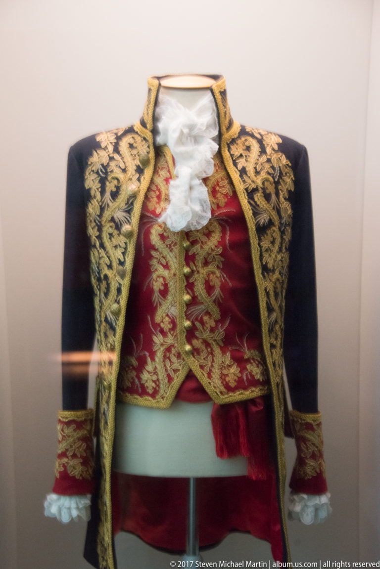 Museum mannequin wearing a dark gold-embroidered jacket, red embroidered waistcoat, white lace, and pale waist sash.