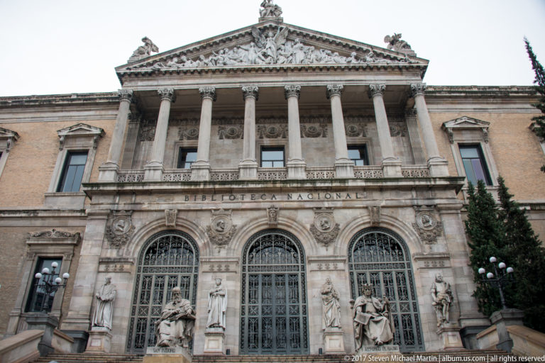 Biblioteca Nacional de España façade with Corinthian columns, arched iron doors, statues, staircase, and sculpted pediment.