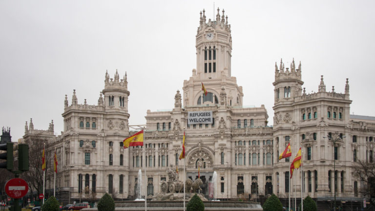 Palacio de Cibeles with Spanish flags and a large “REFUGEES WELCOME” banner hanging from its central façade.
