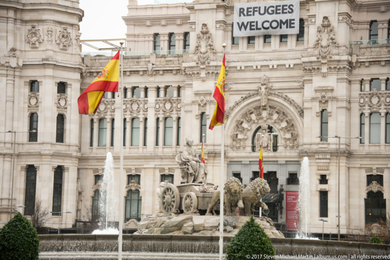 Cibeles Fountain with chariot and lions before the Palacio de Cibeles, Spanish flags, and a “REFUGEES WELCOME” banner.