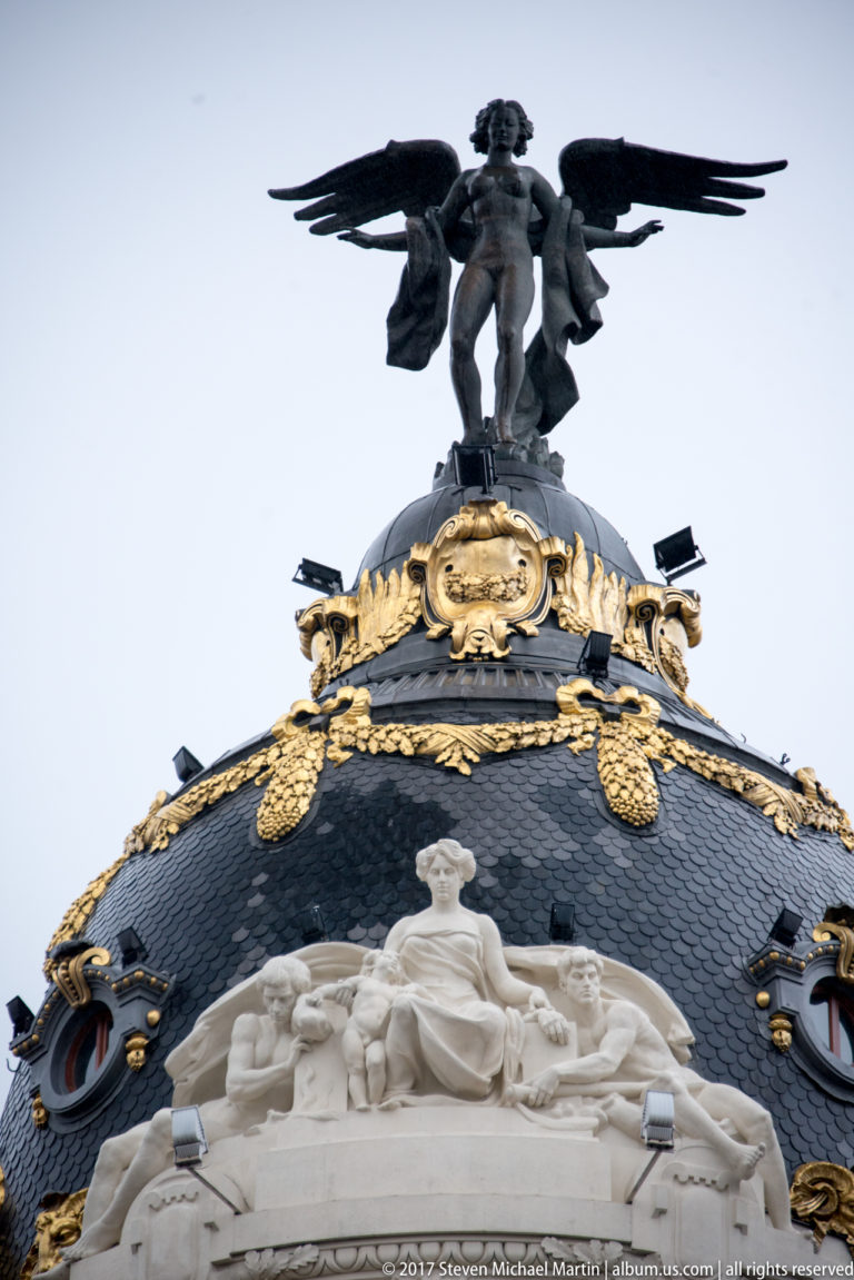 Close view of the Metrópolis Building dome with gilded ornament, seated stone figures, and a winged rooftop sculpture.