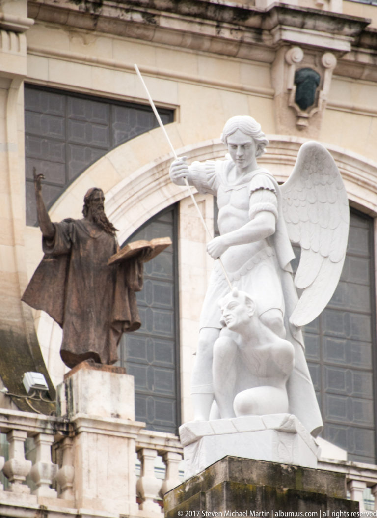 Dark bronze robed figure beside a white winged spear-bearing sculpture above a crouching figure on a palace balustrade.