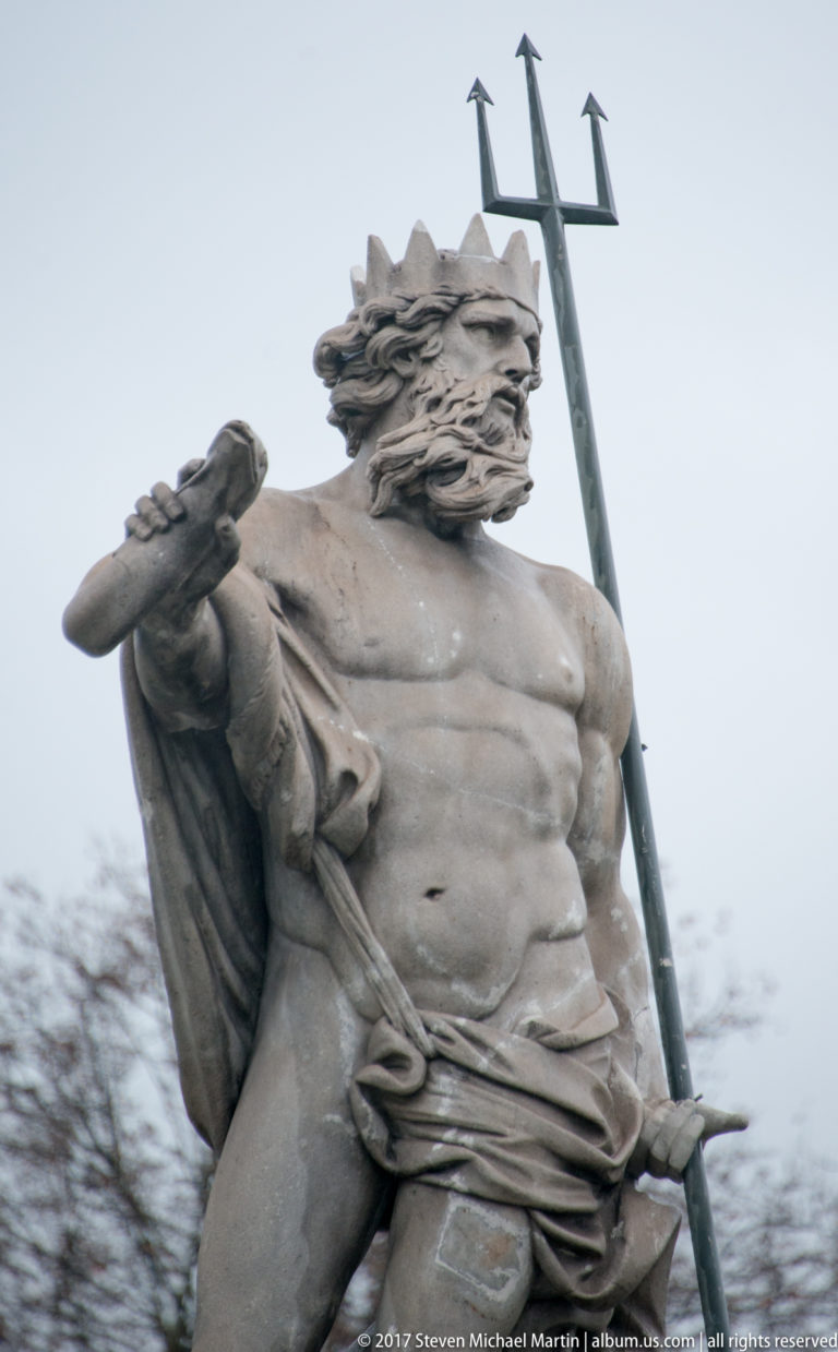 Low-angle close-up of a bearded stone Neptune holding a tall trident against an overcast sky.