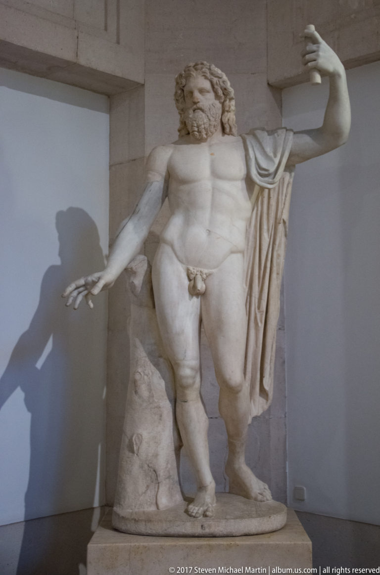 Classical nude male statue with beard, raised arm, drapery, and strong shadow on an interior wall.