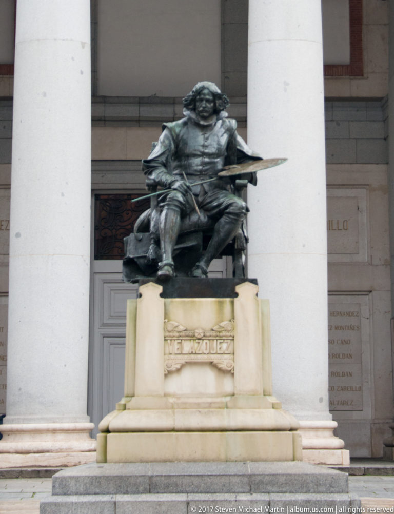 Bronze seated statue of Velázquez holding a palette, on a pedestal inscribed VELAZQUEZ before stone columns.