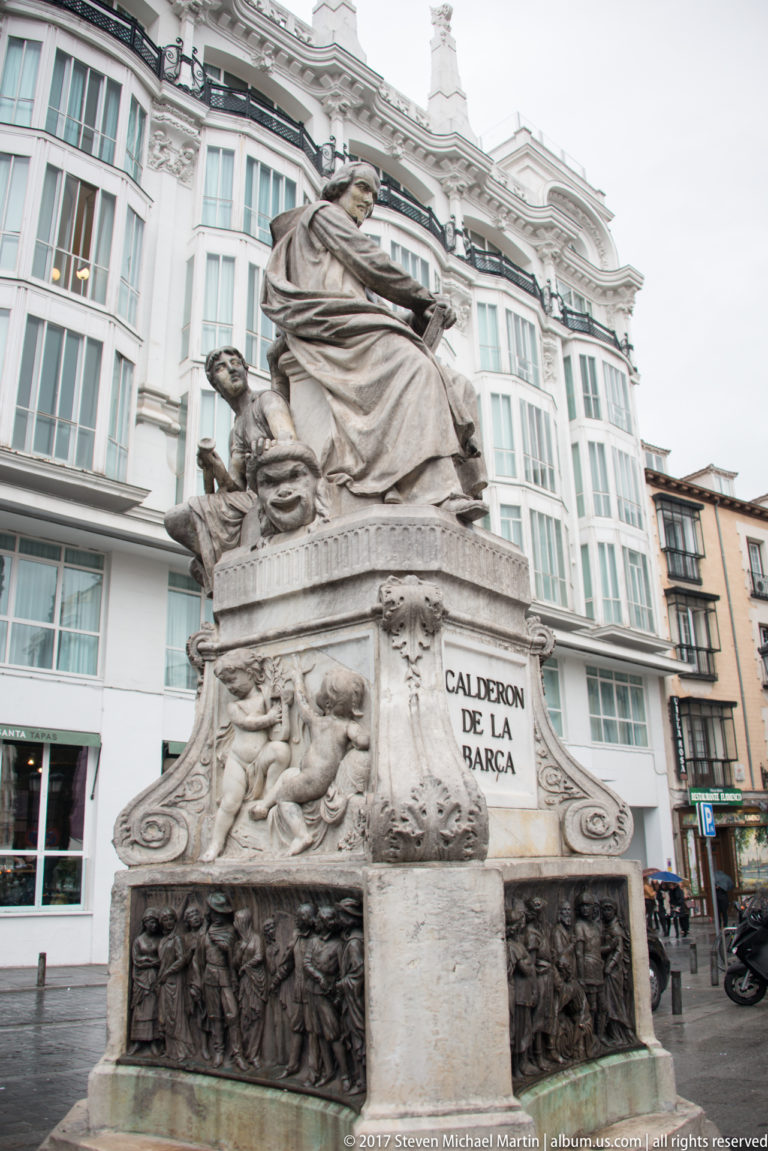 Stone monument to Calderón de la Barca with seated figure, inscribed pedestal, relief panels, and surrounding city buildings.
