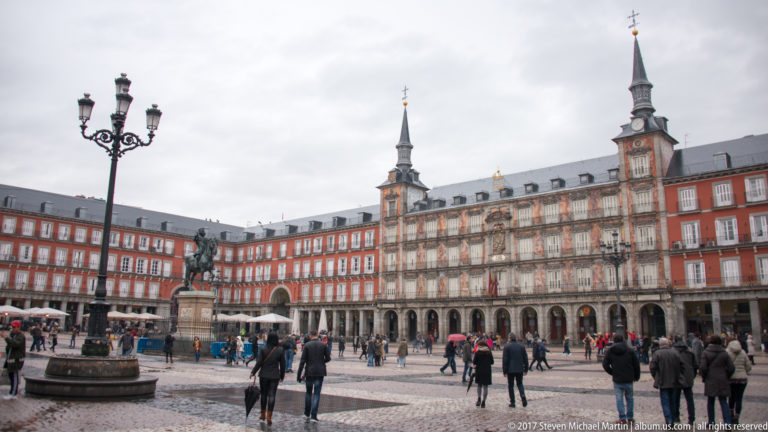 Wide view across Plaza Mayor with red arcaded façades, pedestrians, the equestrian statue of Philip III, and Casa de la Panadería.