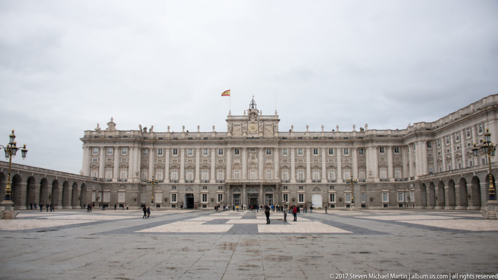 Wide frontal view of Madrid’s Royal Palace across an expansive paved square with Spanish flag and scattered visitors.
