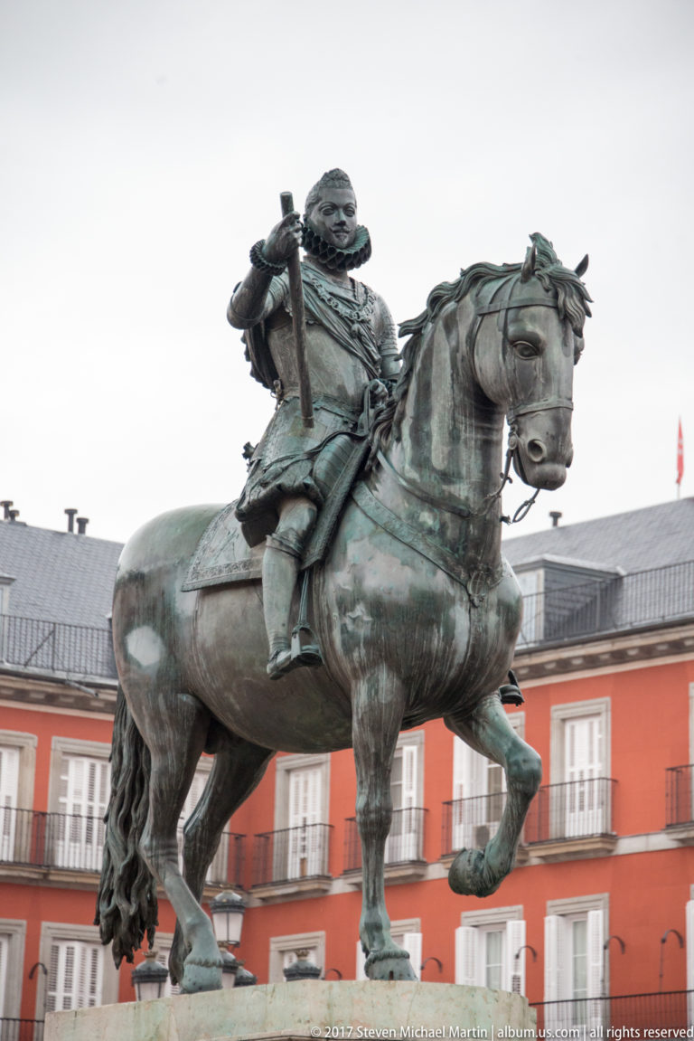 Bronze equestrian statue of Philip III with one horse foreleg raised, set against the red façades of Plaza Mayor.