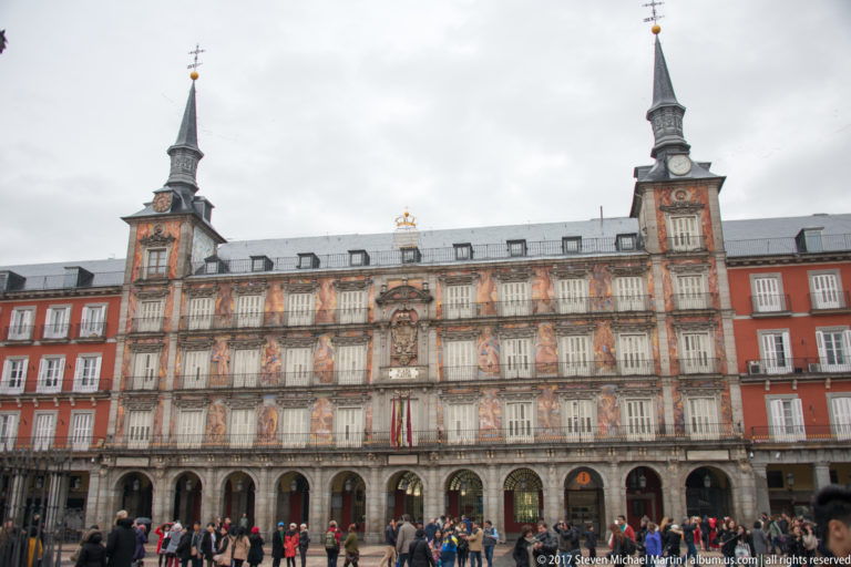 Frontal view of Casa de la Panadería with frescoes, balconies, twin towers, arcades, flags, and visitors in Plaza Mayor.