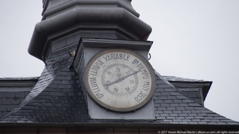 Round Spanish-language weather dial with central pointer mounted on a small slate-clad roof structure.