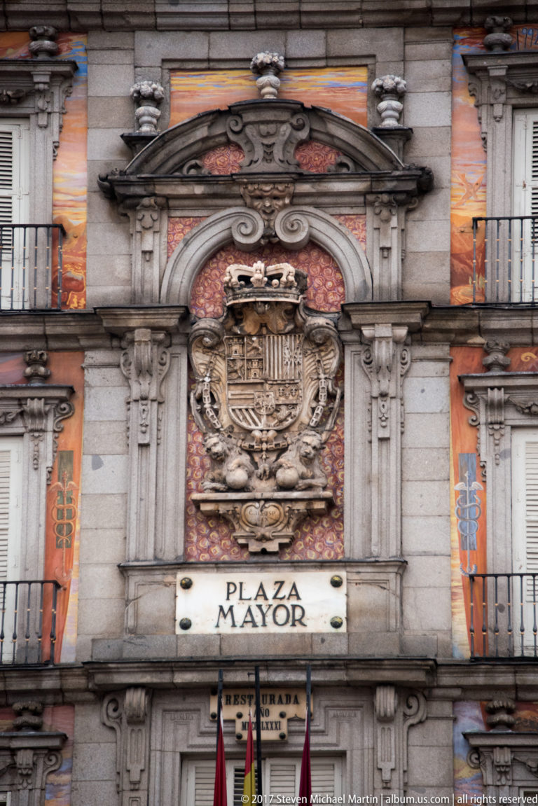 Large crowned coat of arms above a PLAZA MAYOR plaque on an ornate painted and stone façade.