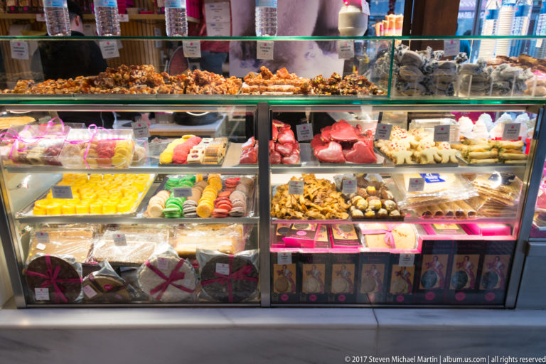 Glass confectionery case filled with colorful macarons, pastries, chocolates, fruit sweets, and labeled trays.