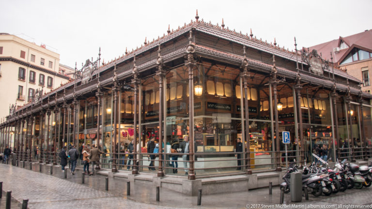 Oblique street view of Mercado de San Miguel with iron columns, glass walls, interior stalls, pedestrians, and parked scooters.