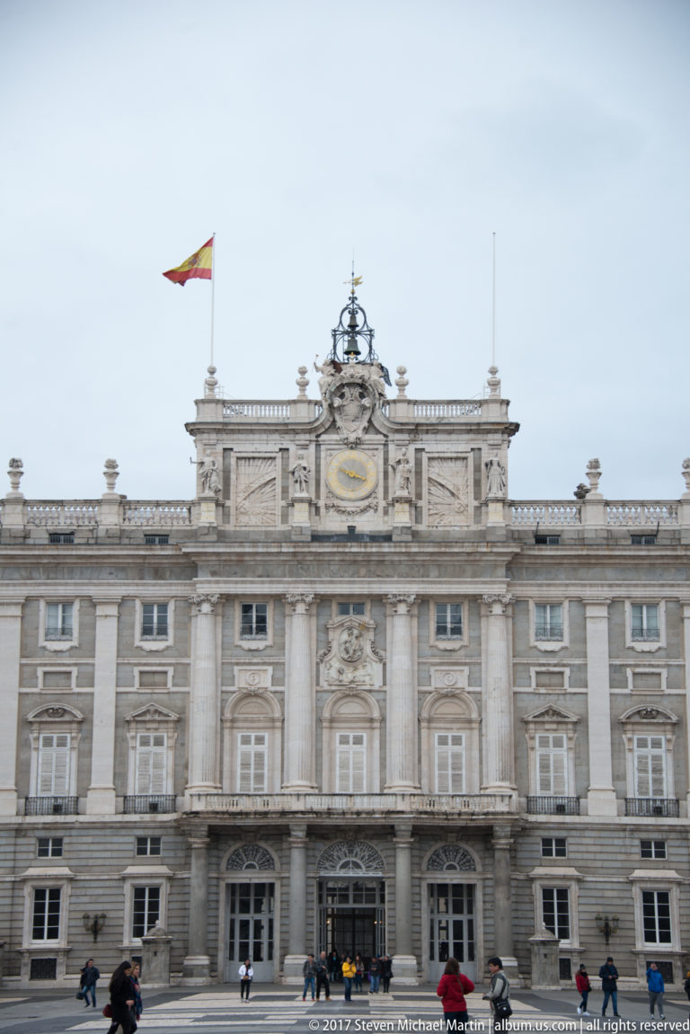 Central section of Madrid’s Royal Palace with clock, sculpted roofline, Spanish flag, and three arched entrance bays.