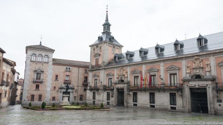 Plaza de la Villa with Casa de la Villa at right, clock tower, flags, stone portals, and adjoining historic buildings.