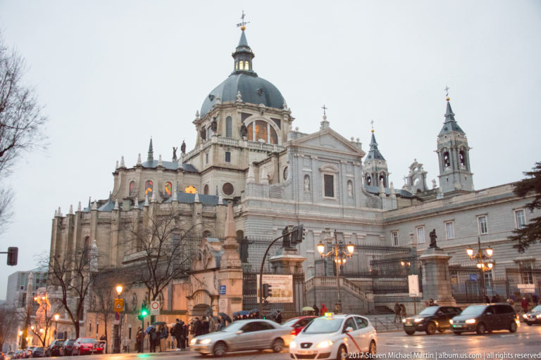 Almudena Cathedral with central dome and towers rising above evening traffic, pedestrians, and illuminated street lamps.