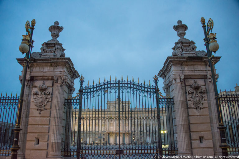 Ornate wrought-iron gates and carved stone piers framing the illuminated Royal Palace of Madrid at dusk.