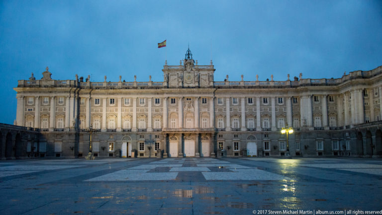 Frontal evening view of the Royal Palace of Madrid with Spanish flag, illuminated windows, and wet plaza pavement.