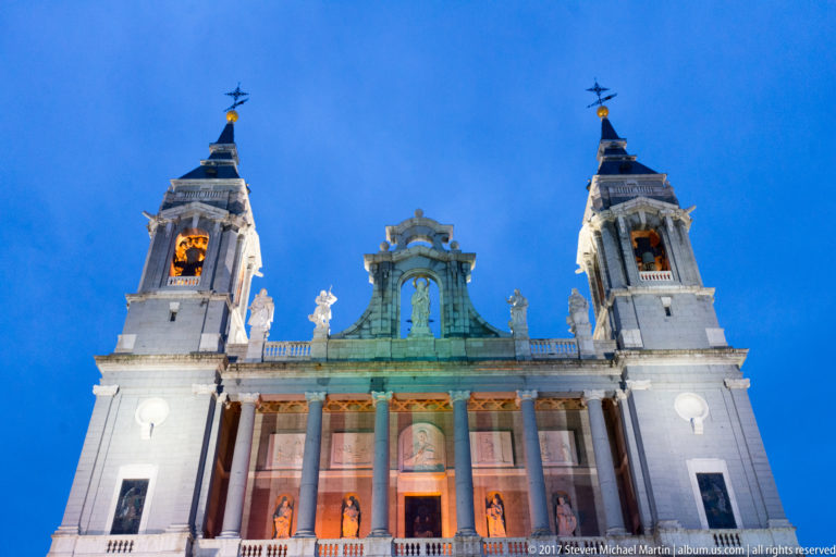 Low-angle evening view of Almudena Cathedral’s illuminated twin towers, colonnaded façade, statues, and central upper opening.
