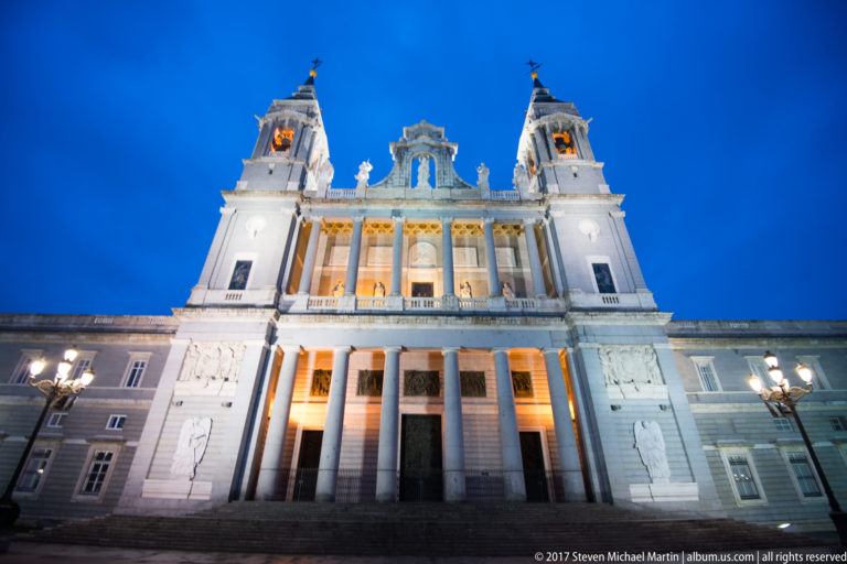 Centered blue-hour view of Almudena Cathedral’s full illuminated façade with twin towers, colonnade, statues, and flanking walls.