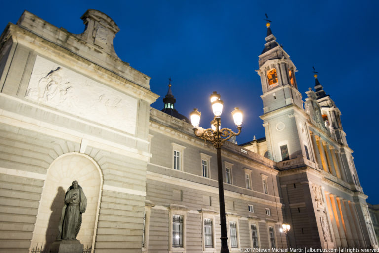 Oblique night view of Almudena Cathedral towers beside pale stone walls, an arched statue niche, and an ornate street lamp.