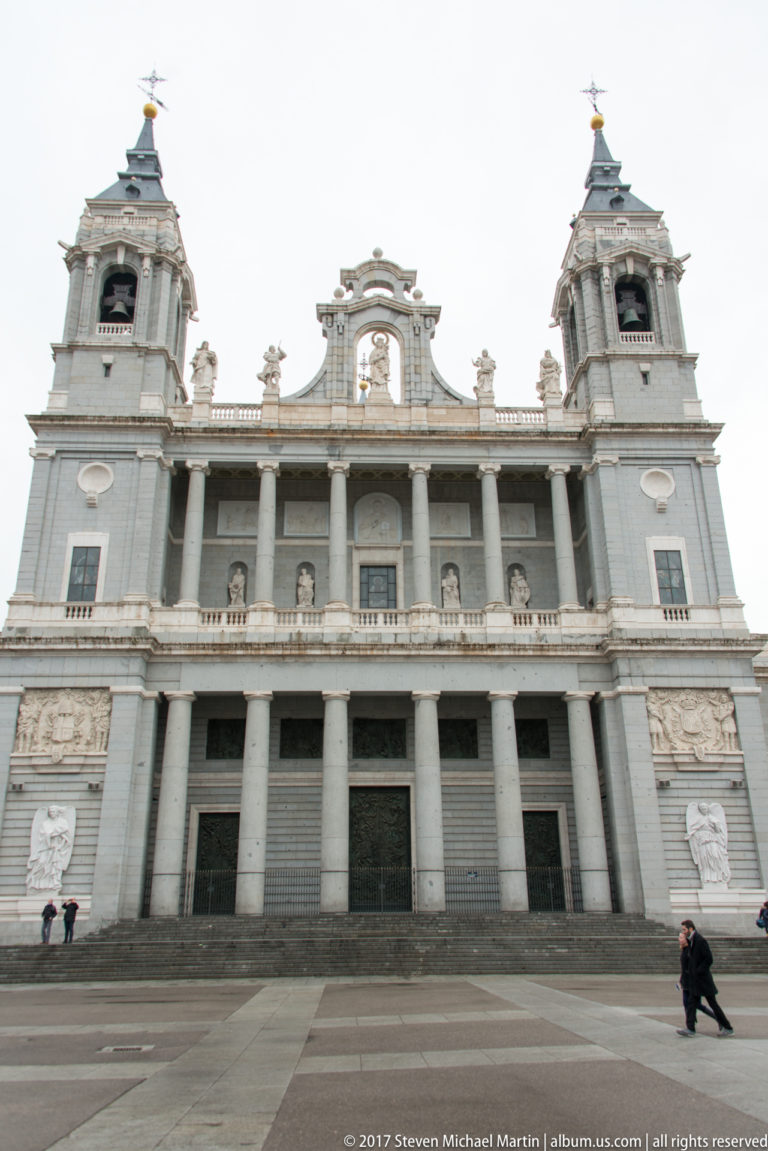 Front façade of Almudena Cathedral with twin towers, central colonnade, broad steps, and pale stone exterior.