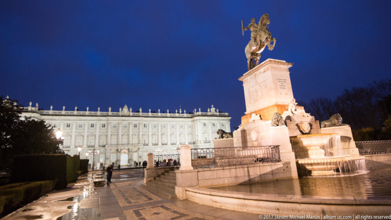 Oblique blue-hour view of the Philip IV monument and fountain with the illuminated Royal Palace extending behind it.