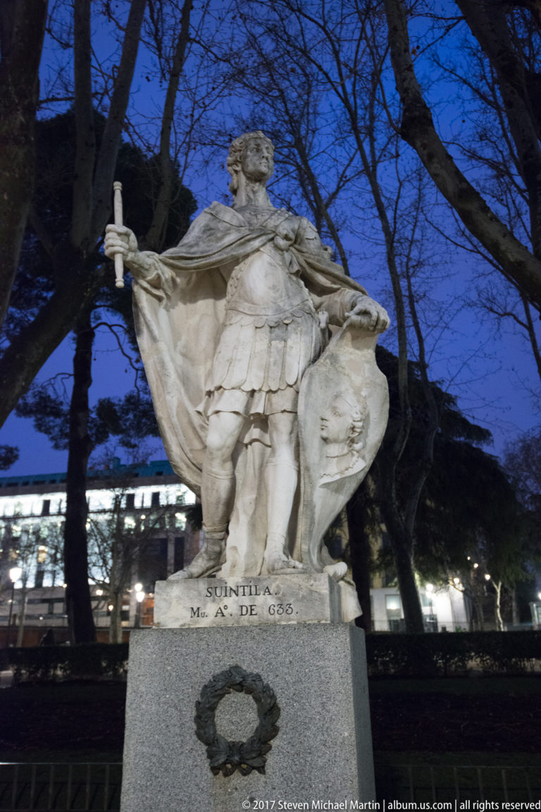 Stone statue of King Suintila in armor and cloak, holding an attribute beside a shield above an inscribed pedestal.