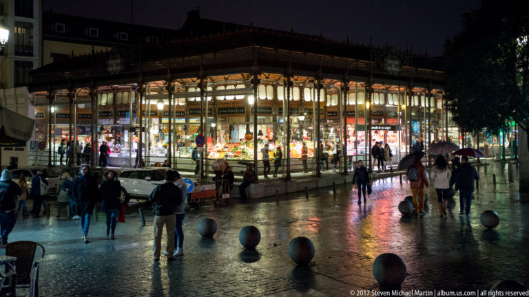Mercado de San Miguel glowing through iron-and-glass walls at night, with pedestrians, umbrellas, and reflections on wet pavement.
