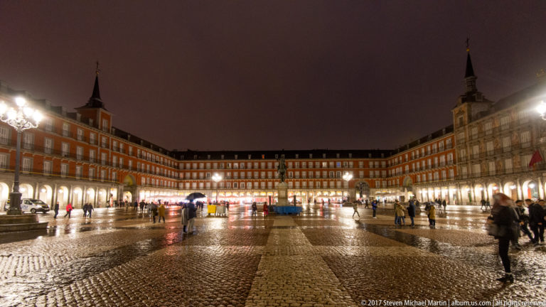 Wide nighttime view across wet Plaza Mayor with red arcaded façades, pedestrians, bright lamps, and the Philip III statue.