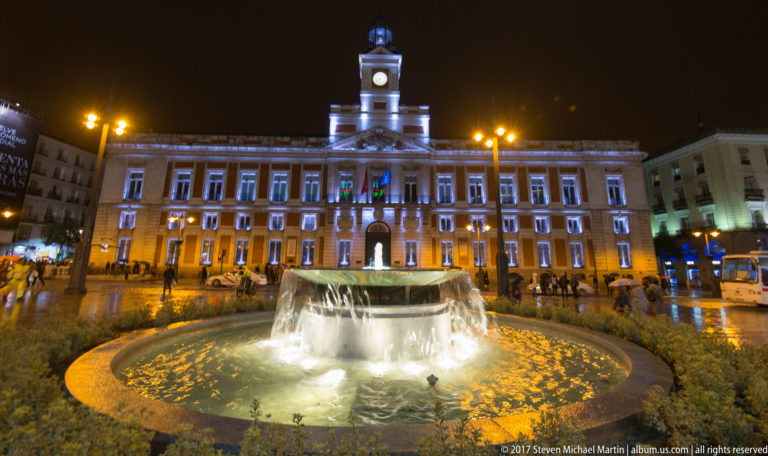 Real Casa de Correos at Puerta del Sol at night, with illuminated clock tower and bright circular fountain in the foreground.