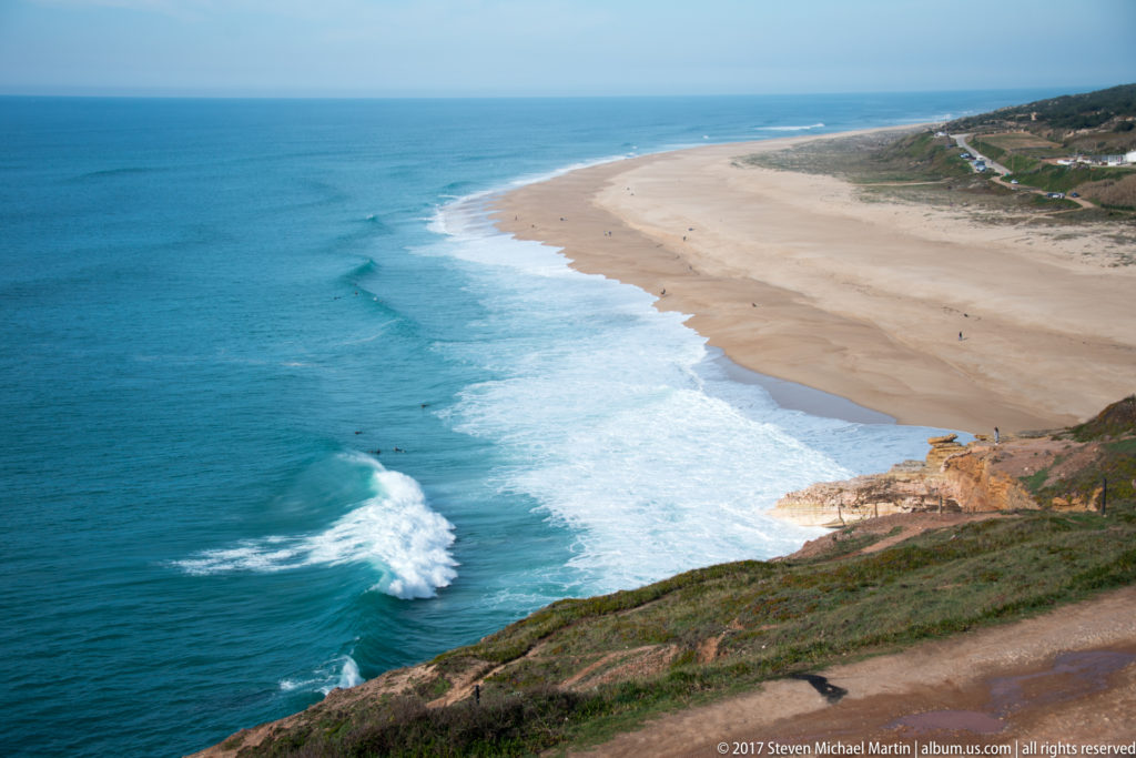 Long sandy Atlantic beach curving north below a cliff, with white surf lines and blue ocean.