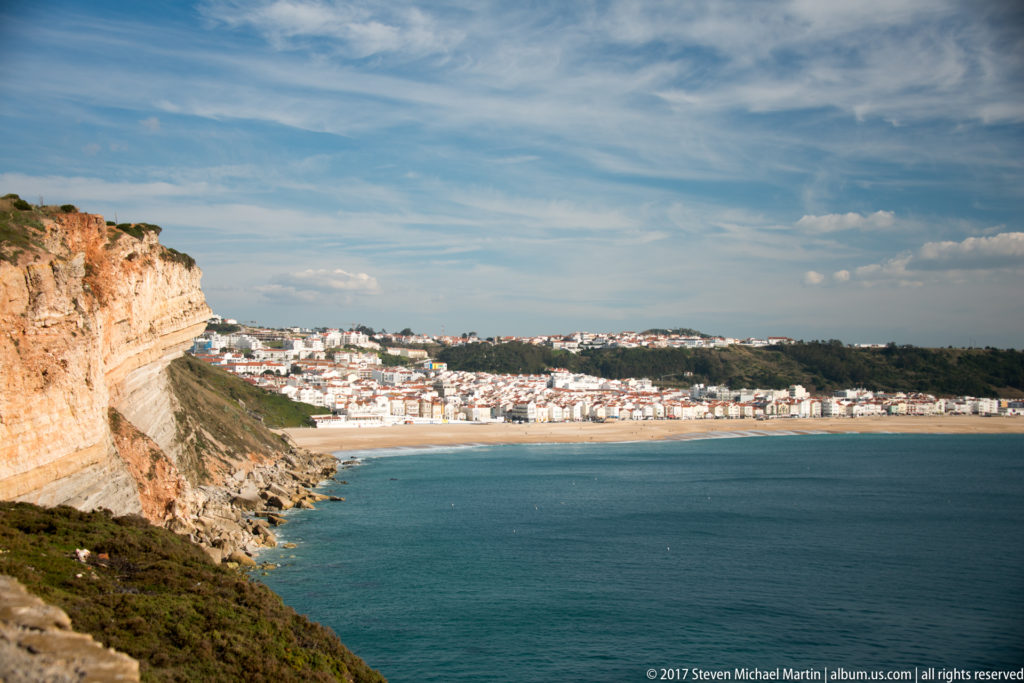 High view past a sheer cliff toward Nazaré’s beach, bay, and dense white-roofed town.