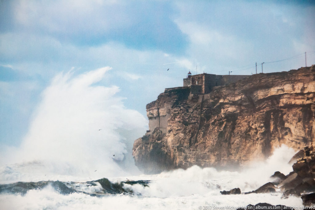 Large wave exploding into white spray against a high cliff with a small lighthouse above.
