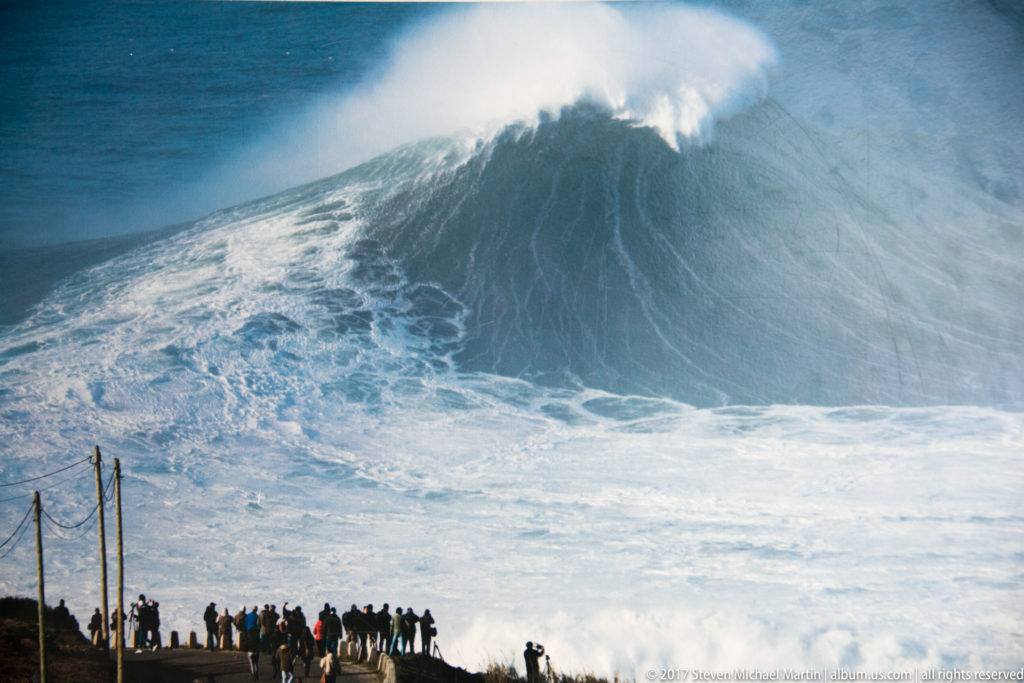 Crowd watching an enormous breaking wave whose crest towers above them in wind-blown spray.