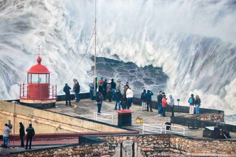 People beside a red lighthouse as an immense white wave breaks directly behind the fort terrace.