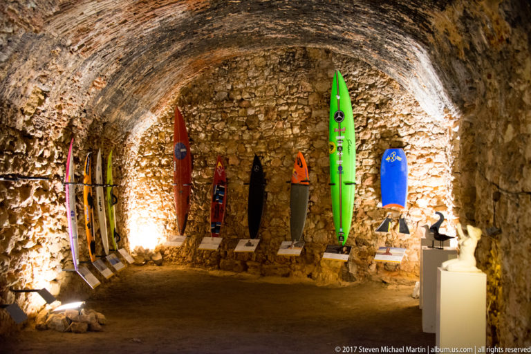 Colorful surfboards displayed upright along the wall of a rough stone-vaulted interior.