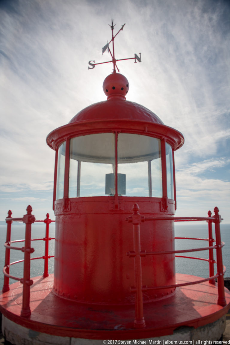 Close frontal view of a red glazed lighthouse lantern, circular gallery, and weather vane.