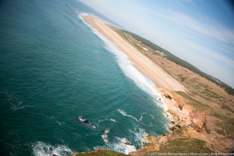 Tilted high-angle view of blue ocean, rocky cliff, and long sandy beach north of Nazaré.