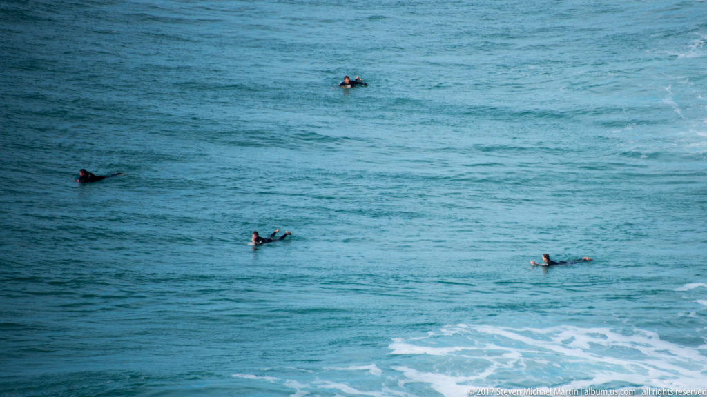Four surfers lying or sitting on boards in blue ocean water off Nazaré.