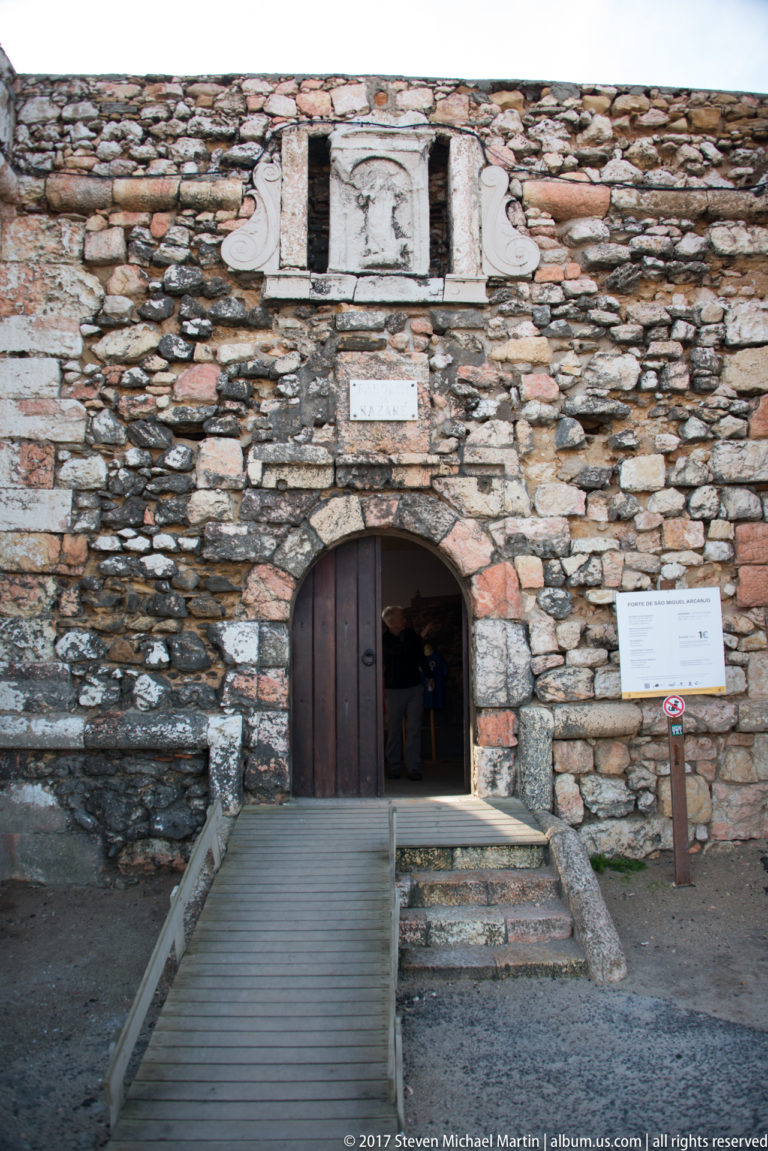Arched wooden doorway in a thick rough-stone fort wall, approached by a narrow ramp.