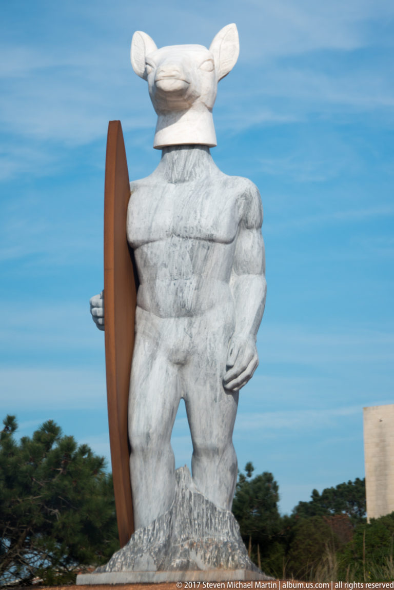 Rear view of a muscular deer-headed stone figure standing beside an upright surfboard.