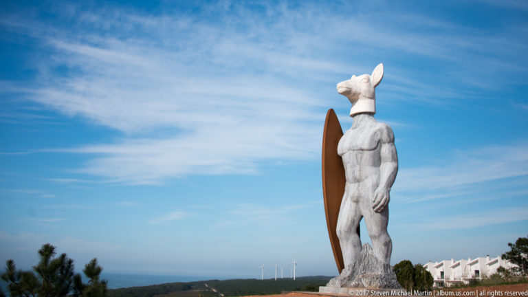 Side view of a deer-headed surfer statue with surfboard against blue sky and distant sea.
