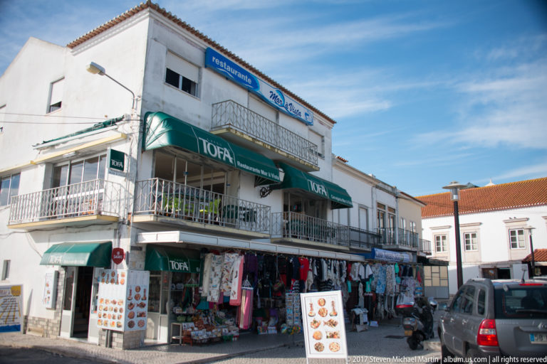 White corner building with balconies, green awnings, souvenir displays, parked cars, and shops in Nazaré.