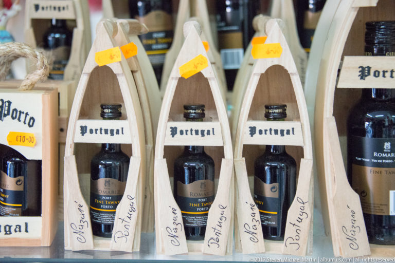 Rows of small Port wine bottles displayed in pale wooden gift holders with handwritten labels and prices.