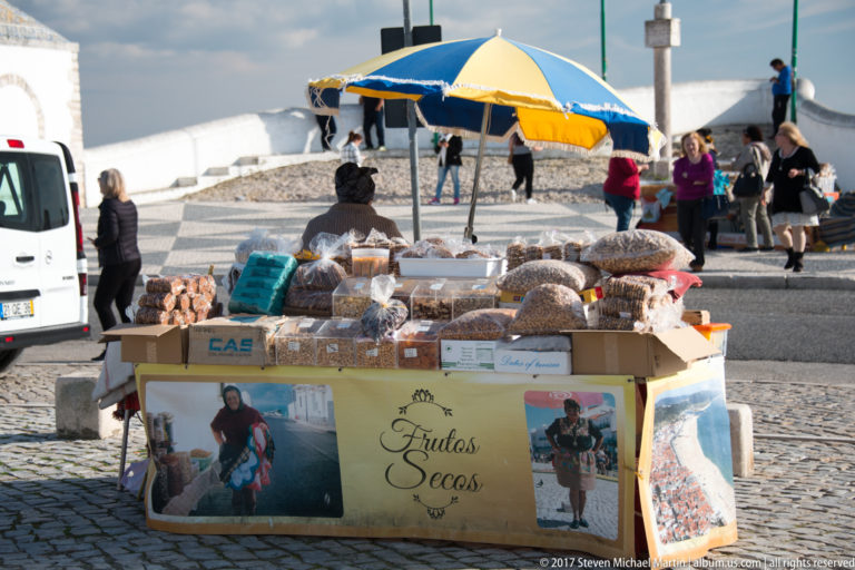 Street cart piled with packaged nuts and dried foods beneath a blue-and-yellow umbrella.