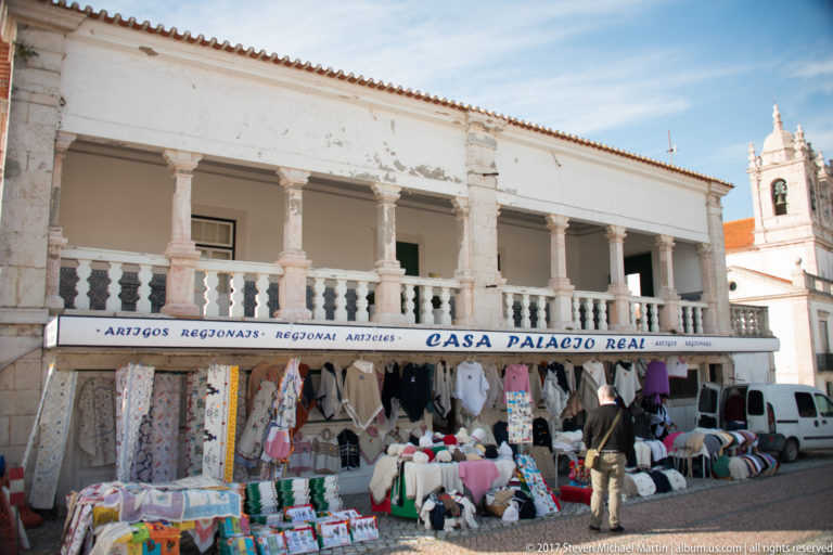 Regional-goods shop beneath a white colonnade, with textiles and hats displayed outside and a church tower behind.