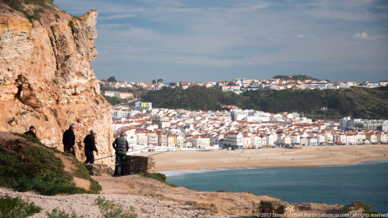 People on a cliffside path overlooking dense white buildings and the beach at Nazaré.